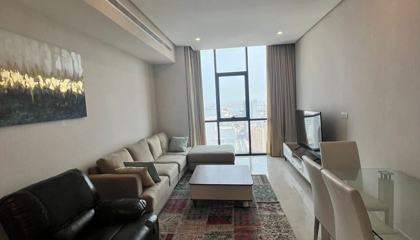 Modern living room with a sofa, armchair, TV, glass dining table, and large window with city view. Neutral tones and contemporary decor throughout.