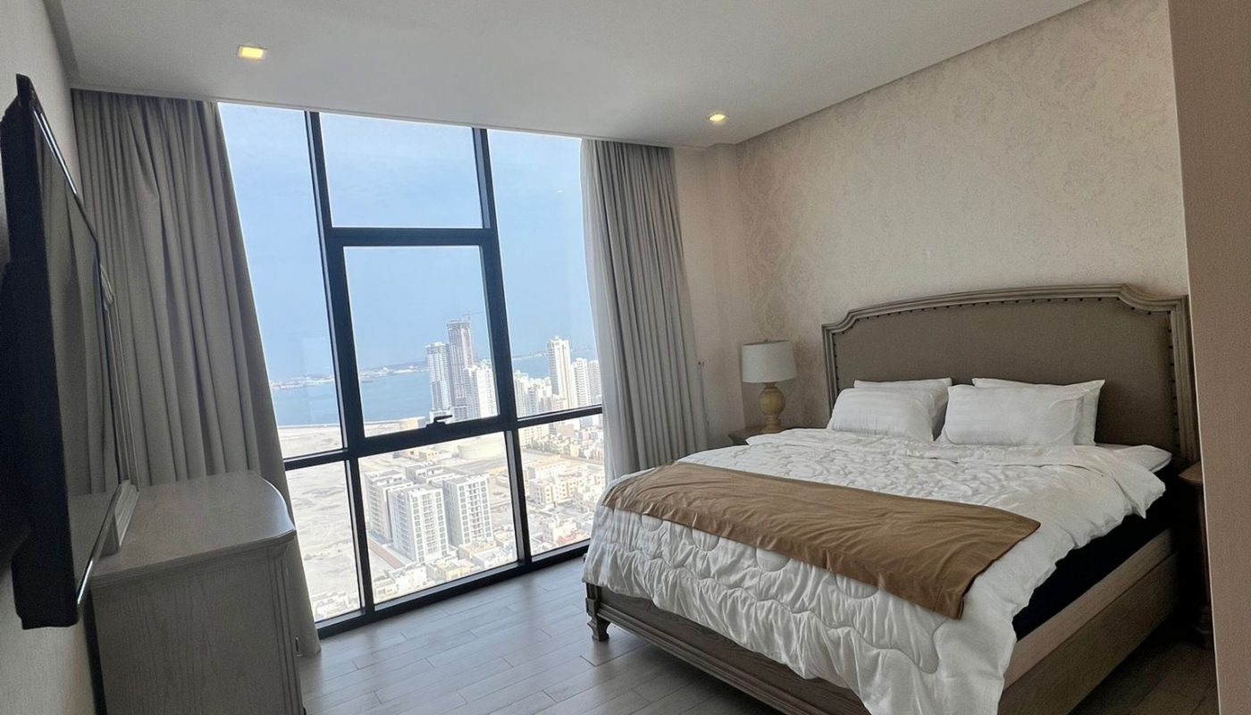 Modern bedroom with a large window offering a city view, featuring a double bed with brown and white bedding, a TV on the wall, and light wood flooring.