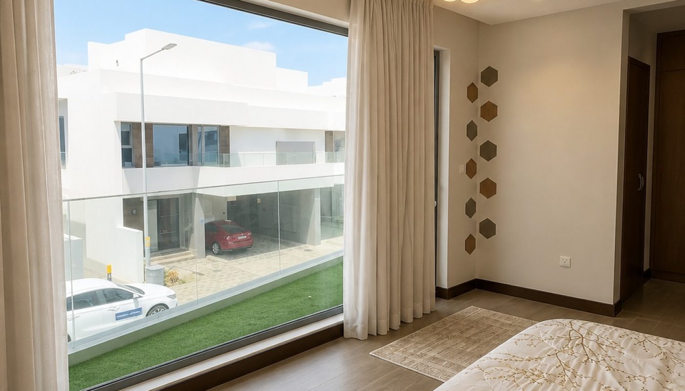 Modern bedroom with large window, neutral decor, patterned bedding, wooden floor, ceiling light fixture, and a view of contemporary houses and parked cars outside.