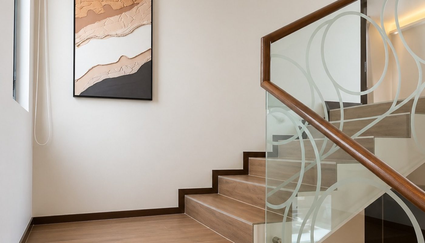 Modern wooden staircase with a glass railing featuring a circular pattern, angled upward toward a wall-mounted abstract artwork and a small wall light.