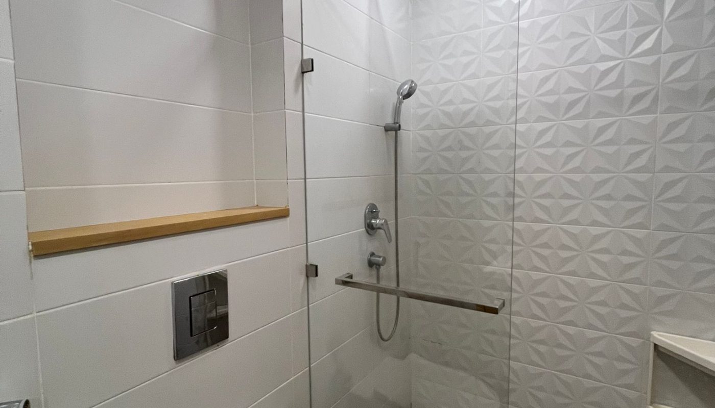 Modern bathroom with a glass-enclosed shower, wall-mounted toilet, white textured tiles, and built-in shelf. Overhead and handheld showerheads are visible.