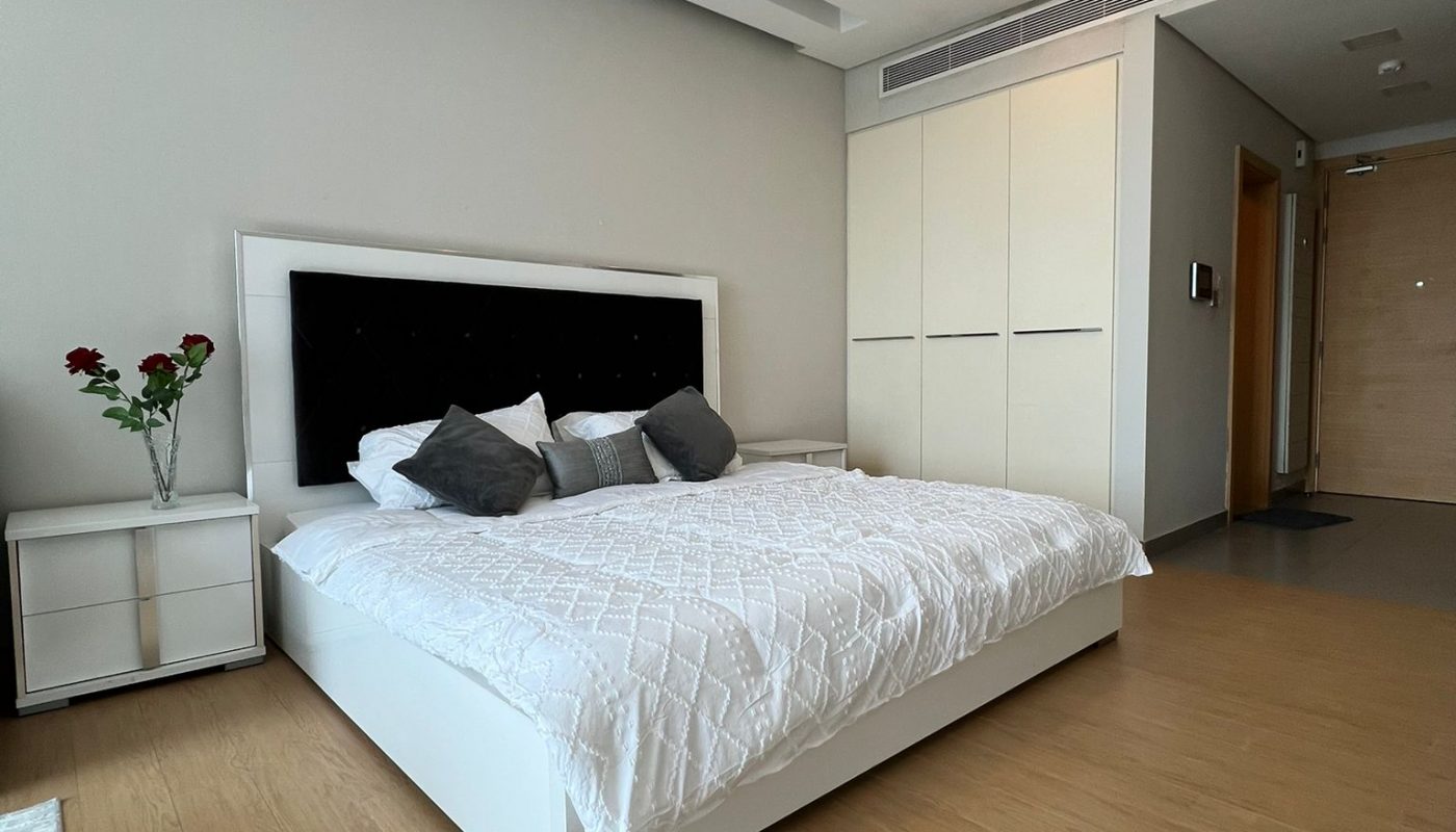 Modern bedroom with a large white bed, white quilt, gray pillows, nightstand with flowers, built-in wardrobe, wood floor, and recessed ceiling lights.