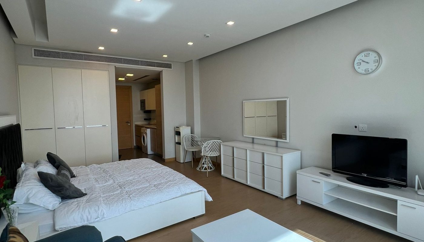 Modern studio apartment with a bed, sofa, coffee table, TV, dresser, and kitchenette. White walls, wooden floors, and minimal decor are visible.