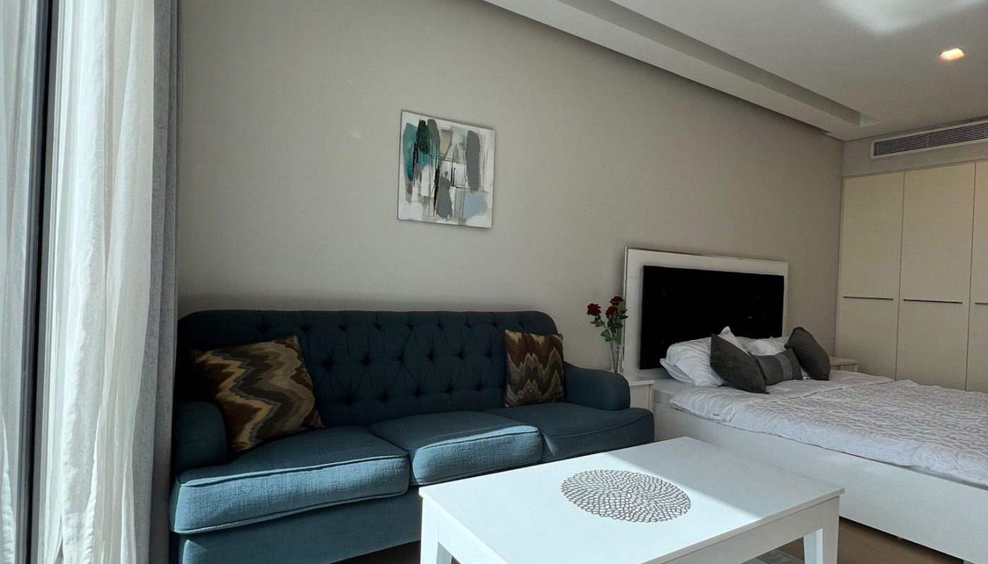 A modern bedroom with a blue sofa, white coffee table, bed with white bedding, abstract wall art, and large windows letting in natural light.