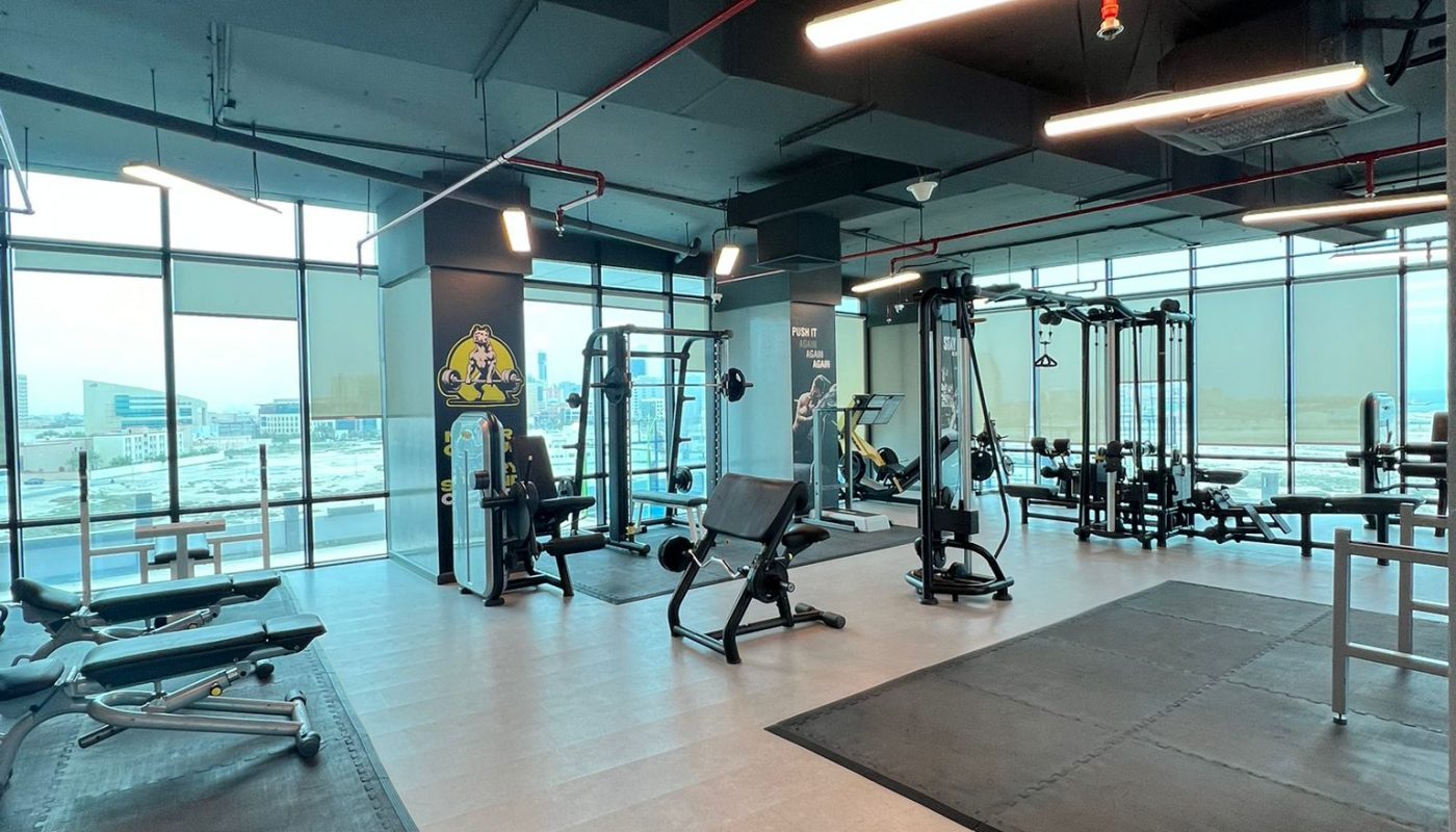 A modern gym with weight machines, benches, and large windows letting in natural light. The space is clean and organized with rubber flooring in workout areas.