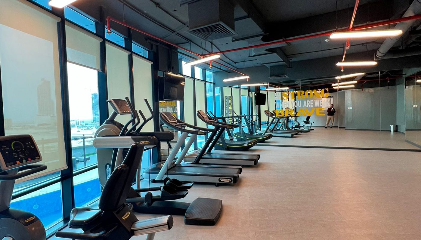 A modern gym with cardio machines including exercise bikes and treadmills, large windows, and bright overhead lighting.