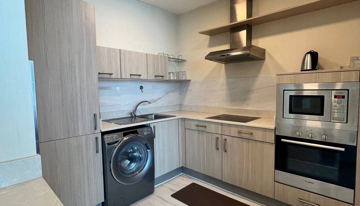 Modern kitchen with light wood cabinets, built-in oven, microwave, washing machine, electric cooktop, sink, and vent hood. Countertops and floor are light-colored.