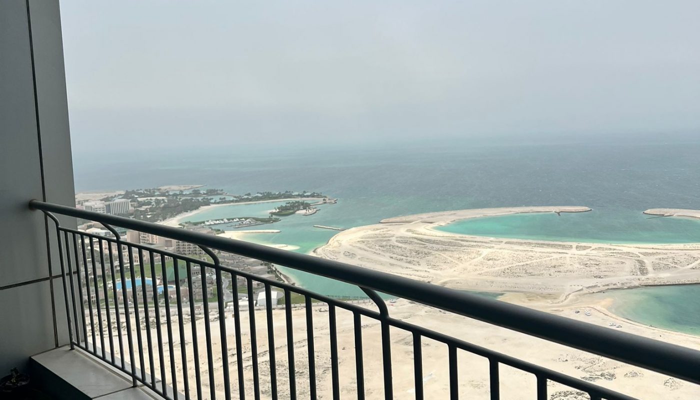 View from a high-rise balcony overlooking sandy islands and turquoise water under a cloudy sky, with part of the building and balcony railing in the foreground.