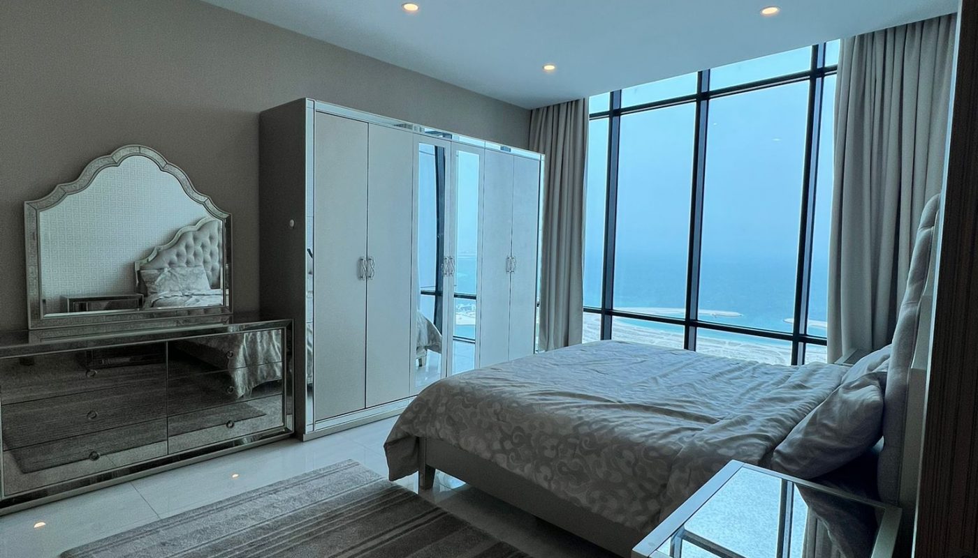 Modern bedroom with a large bed, mirrored wardrobe, dresser, and floor-to-ceiling windows overlooking a body of water. A gray striped rug covers part of the tile floor.