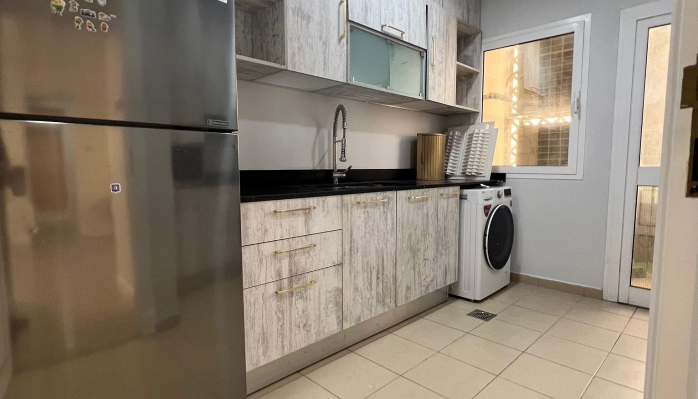 Modern kitchen with light wood cabinets, stainless steel fridge, built-in sink, black countertop, washing machine, and a window letting in natural light.
