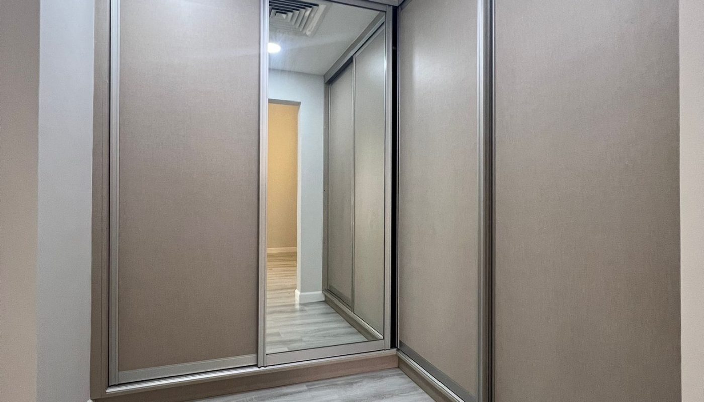 Empty walk-in closet with light wood flooring, beige paneled walls, a ceiling vent, overhead lighting, and a large mirrored sliding door.