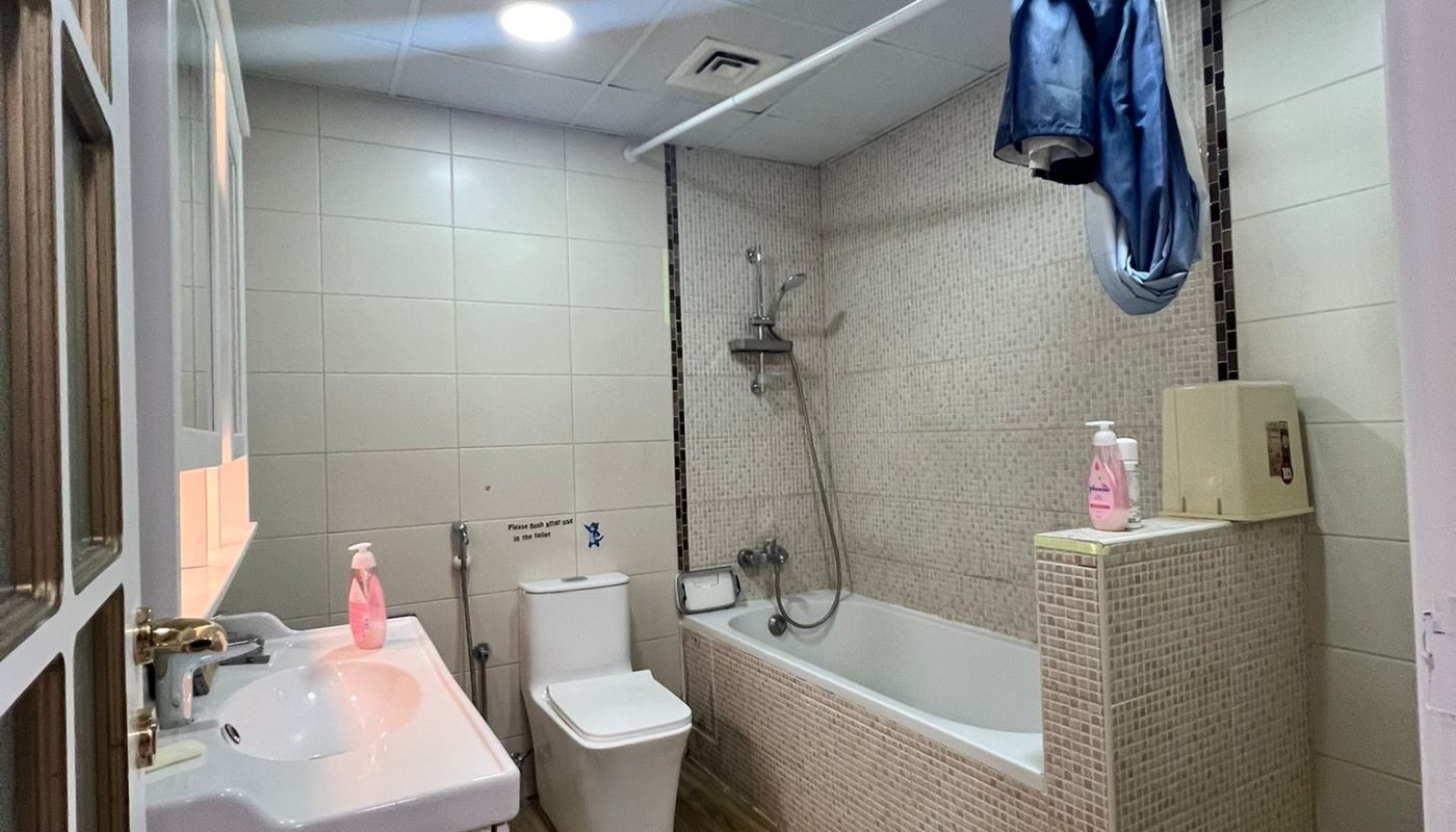 Modern bathroom with a wooden floor, white sink, toilet, bathtub with shower, and a jacket hanging on a pipe above the tub; pink soap bottles on the sink and tub edge.
