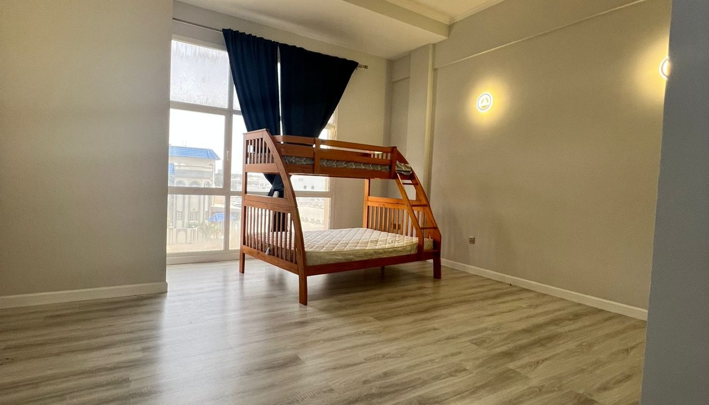 A mostly empty room with light wood flooring, gray walls, and a wooden bunk bed placed near a window with dark curtains. Two wall-mounted lights are visible.