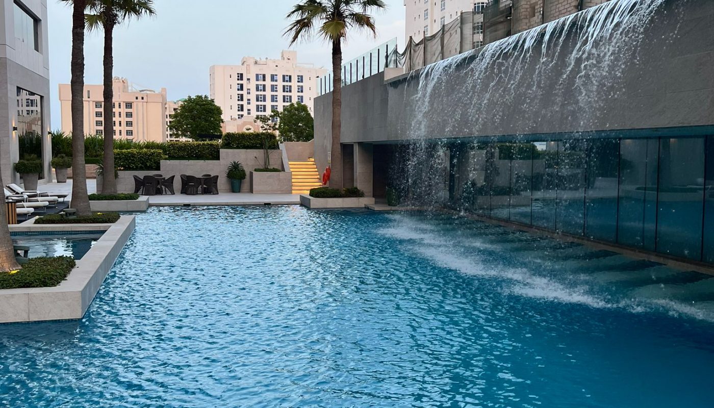A swimming pool with a waterfall feature next to a building under construction, surrounded by palm trees and city buildings in the background.