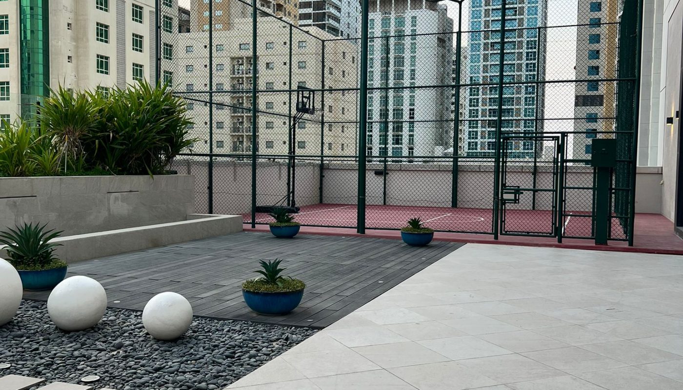Outdoor sports court surrounded by a tall fence, with potted plants and modern high-rise buildings in the background. Concrete and stone walkway in the foreground.