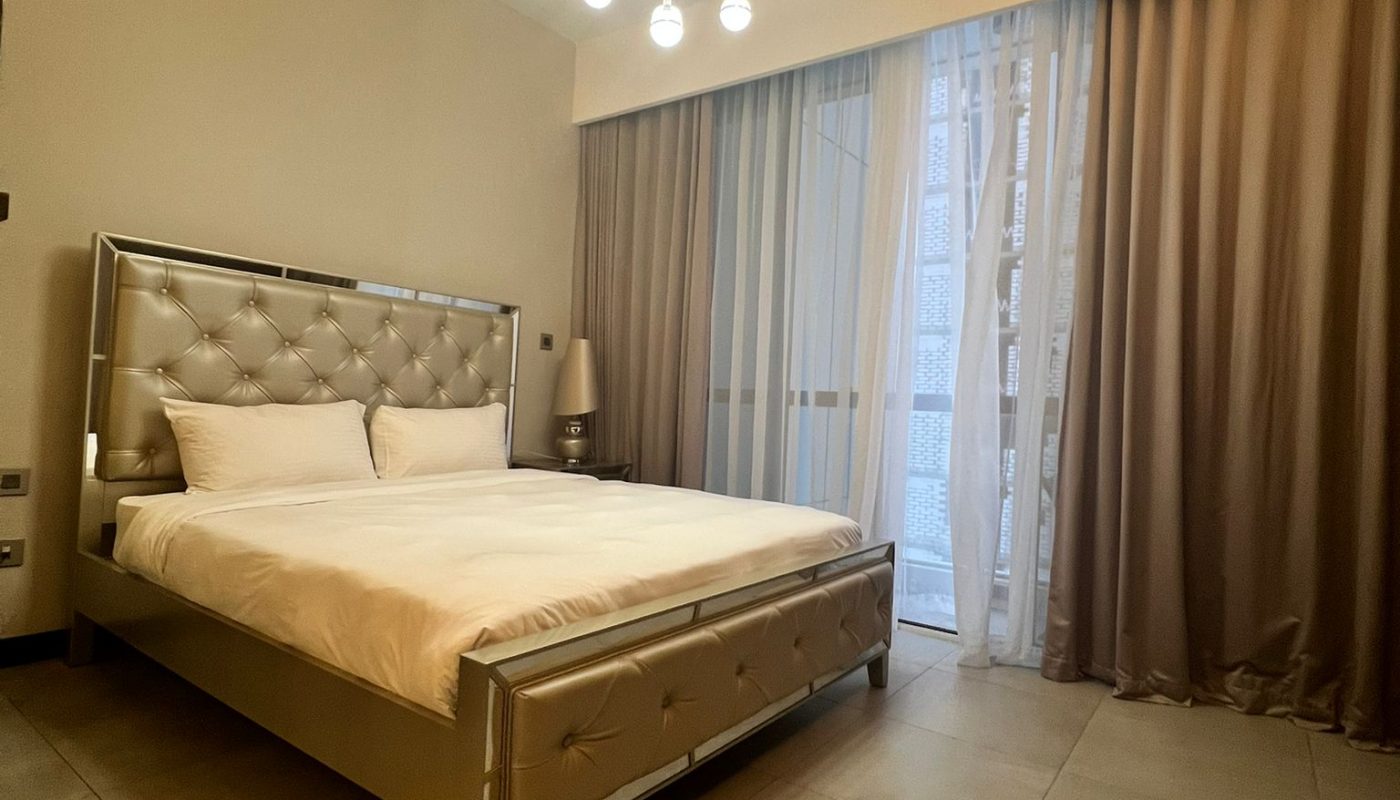 Modern bedroom with a large bed, tufted headboard, neutral bedding, tiled floor, floor-to-ceiling curtains, and a ceiling light fixture with multiple hanging bulbs.