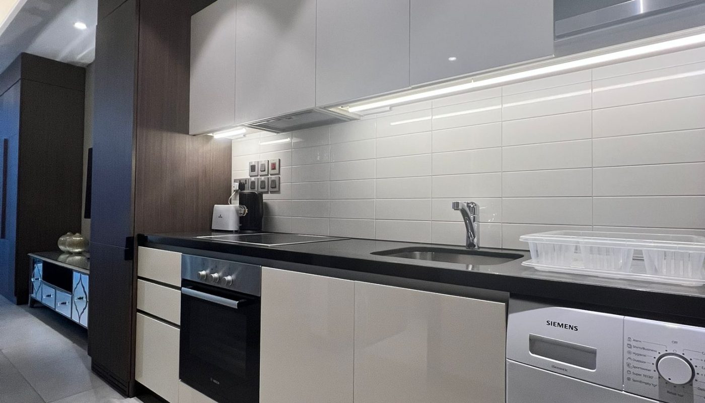 Modern kitchen with white cabinets, built-in oven, washing machine, electric stove, sink, microwave, and tiled backsplash under bright ceiling lights.