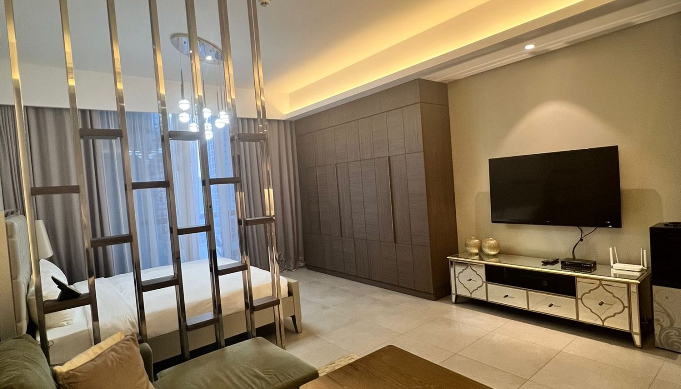Modern hotel suite with a sectional sofa, wooden coffee table, wall-mounted TV, partition separating a bed area, and soft overhead lighting.