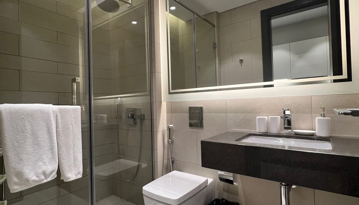 Modern bathroom with a glass shower enclosure, wall-mounted toilet, large mirror, sink with toiletries, towel rack, and a waste bin on beige tiled floor.