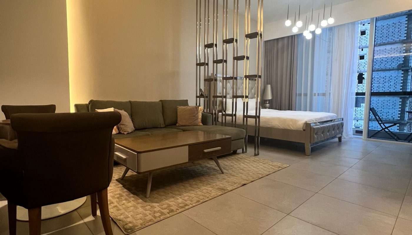 Modern hotel room with a sofa, coffee table, and bed separated by metal rods, large floor tiles, and a window with city views.
