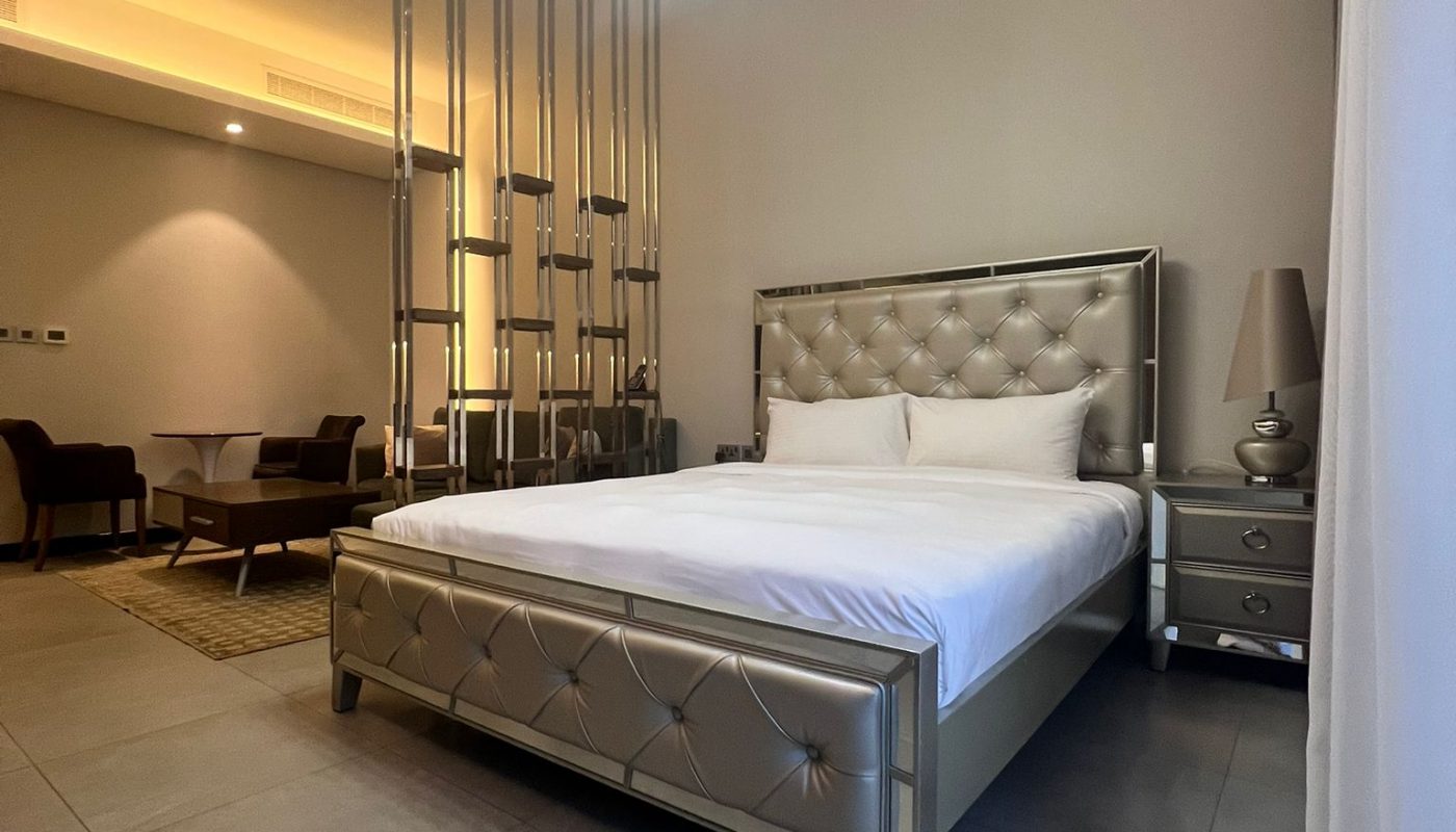 A modern bedroom with a metallic upholstered bed, two nightstands with lamps, a partition with vertical rods, and a seating area in the background.