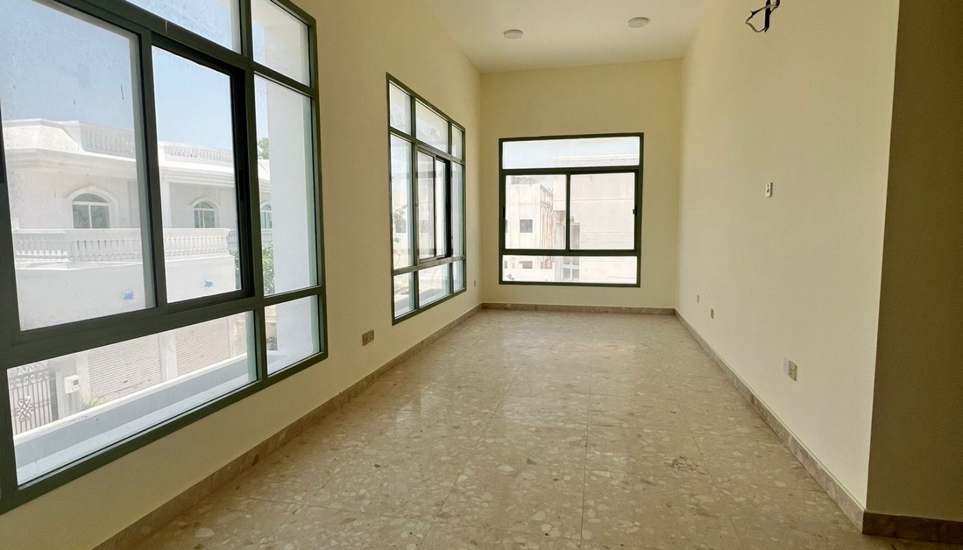 Empty, unfurnished room with large windows on two sides, beige tiled floor, cream-colored walls, and ceiling lights. Daylight is streaming in through the windows.