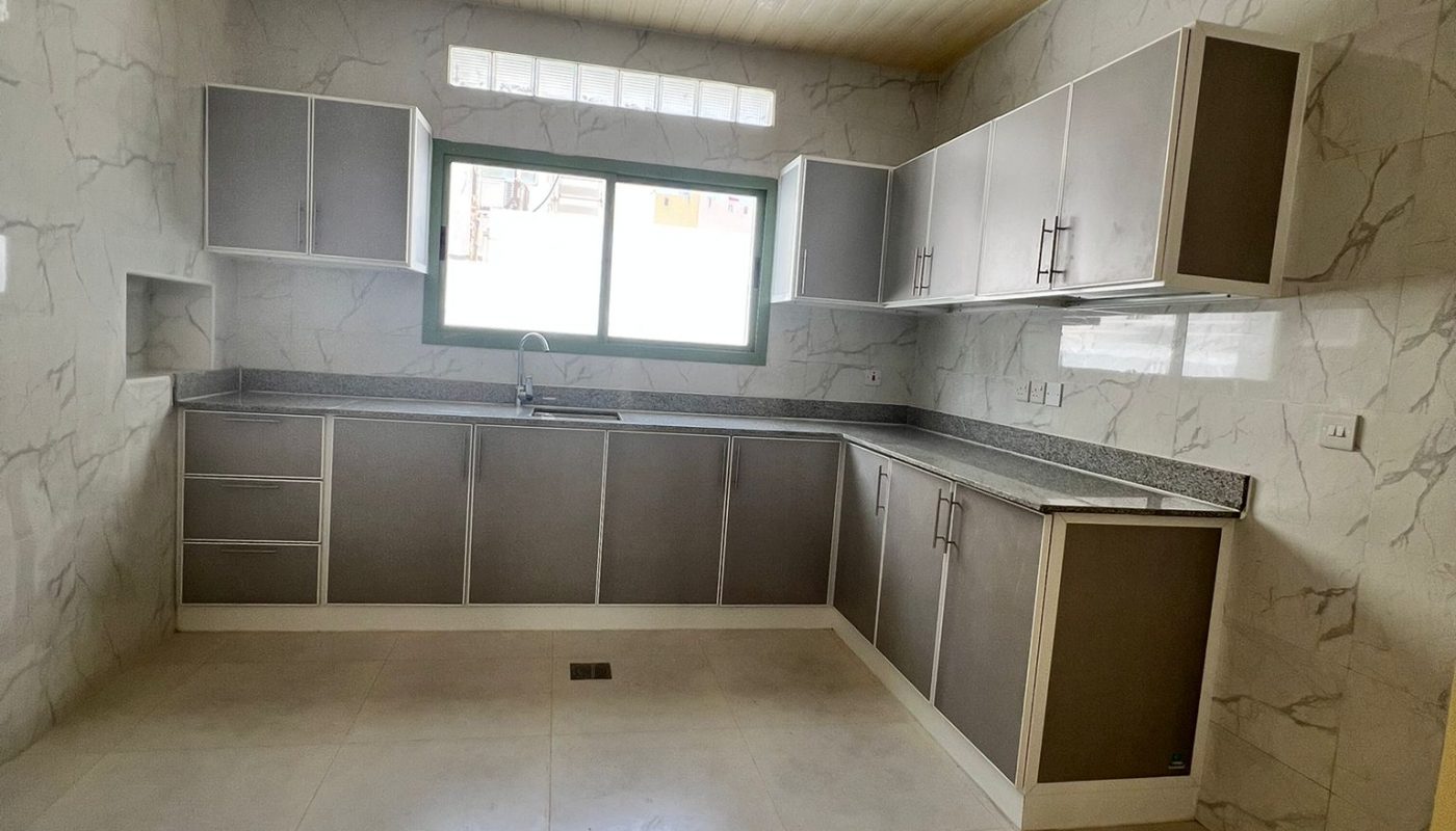 Modern kitchen with marble-patterned walls, beige tiled floor, gray cabinets, a large window above the sink, and recessed ceiling lights.