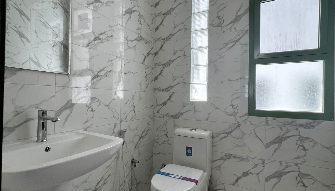 A small bathroom with white marble-patterned tiles, a wall-mounted sink, a toilet, a bidet spray, a mirror, and a window with frosted glass panes.