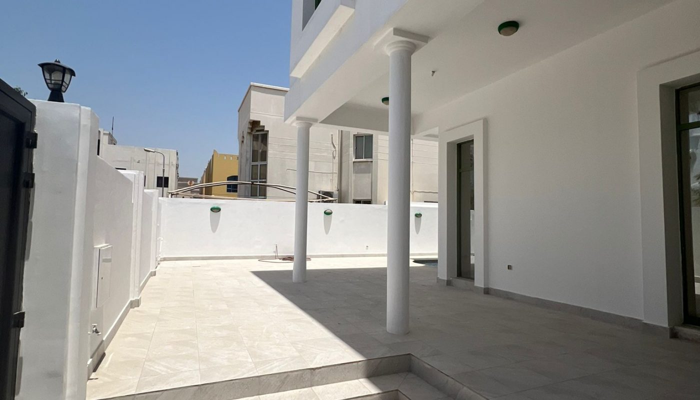 A modern, white two-story house with tiled patio, columns, and a high wall enclosing a sunlit outdoor area.