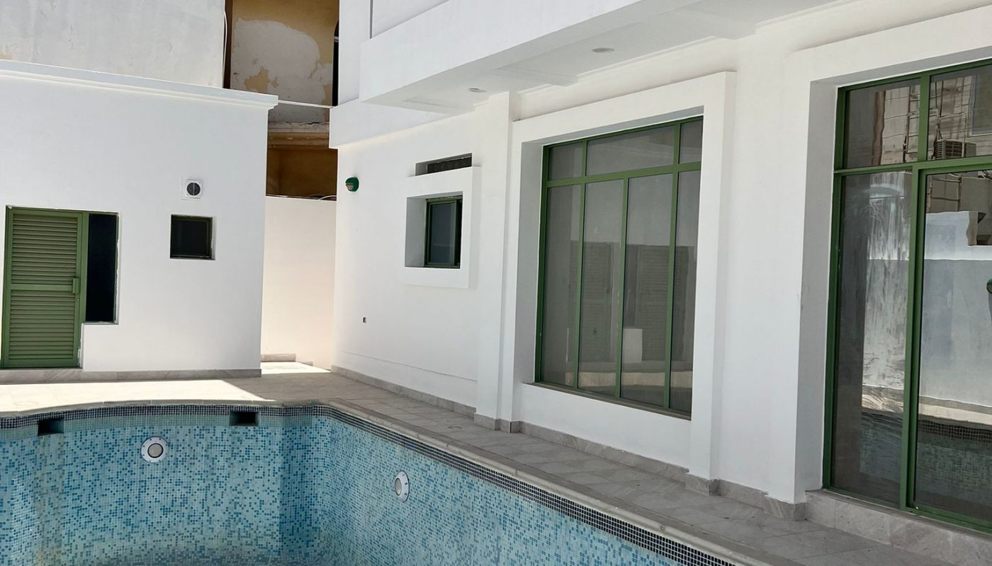 A modern white building with large windows and a small empty swimming pool with blue tiles in a sunny outdoor courtyard.