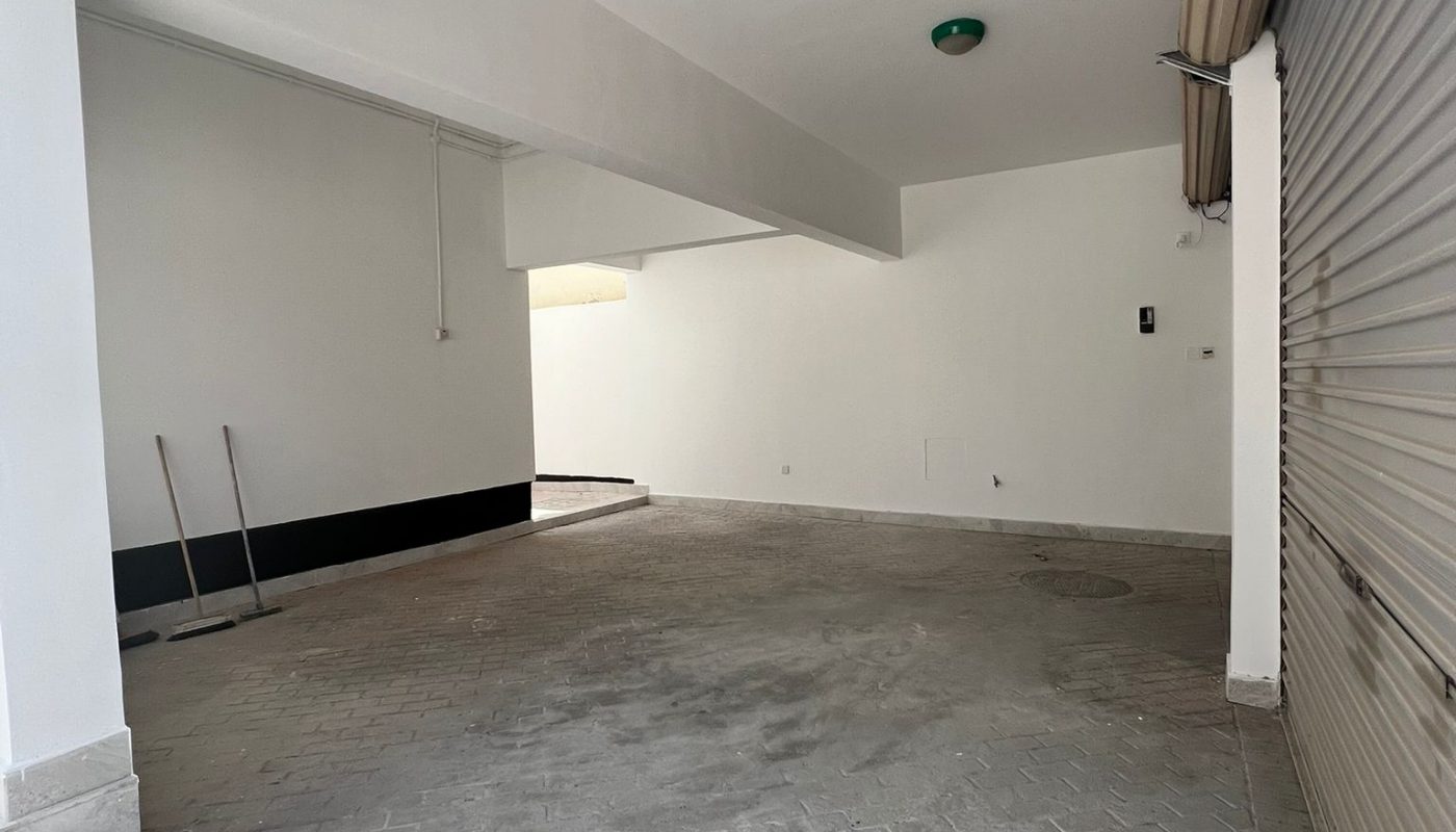 Empty indoor garage with tiled floor, white walls, partially open metal rolling door, and cleaning tools leaning against the wall and lying on the floor.