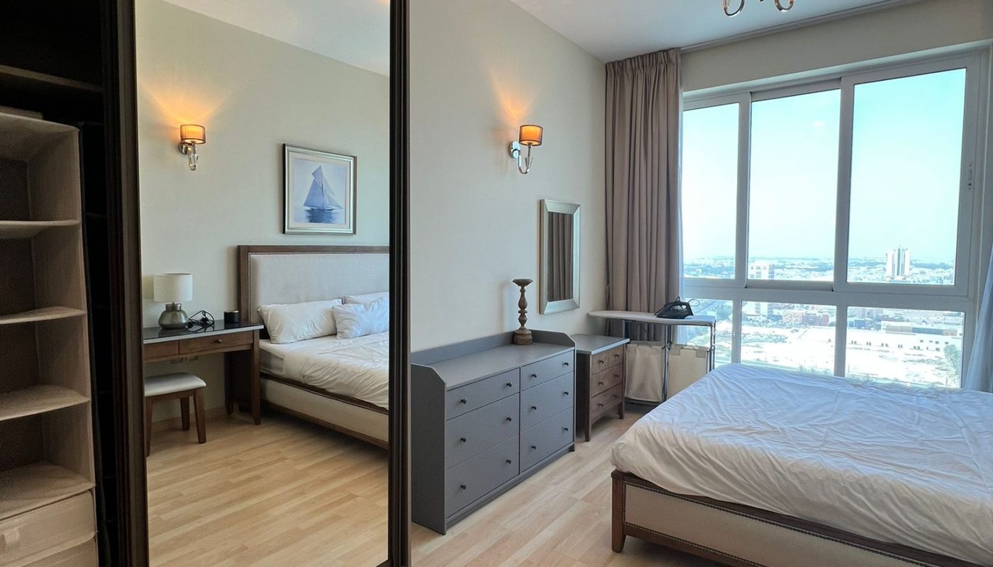 A modern bedroom with a bed, dresser, desk, large mirror wardrobe, light wood flooring, and a window with a city view.