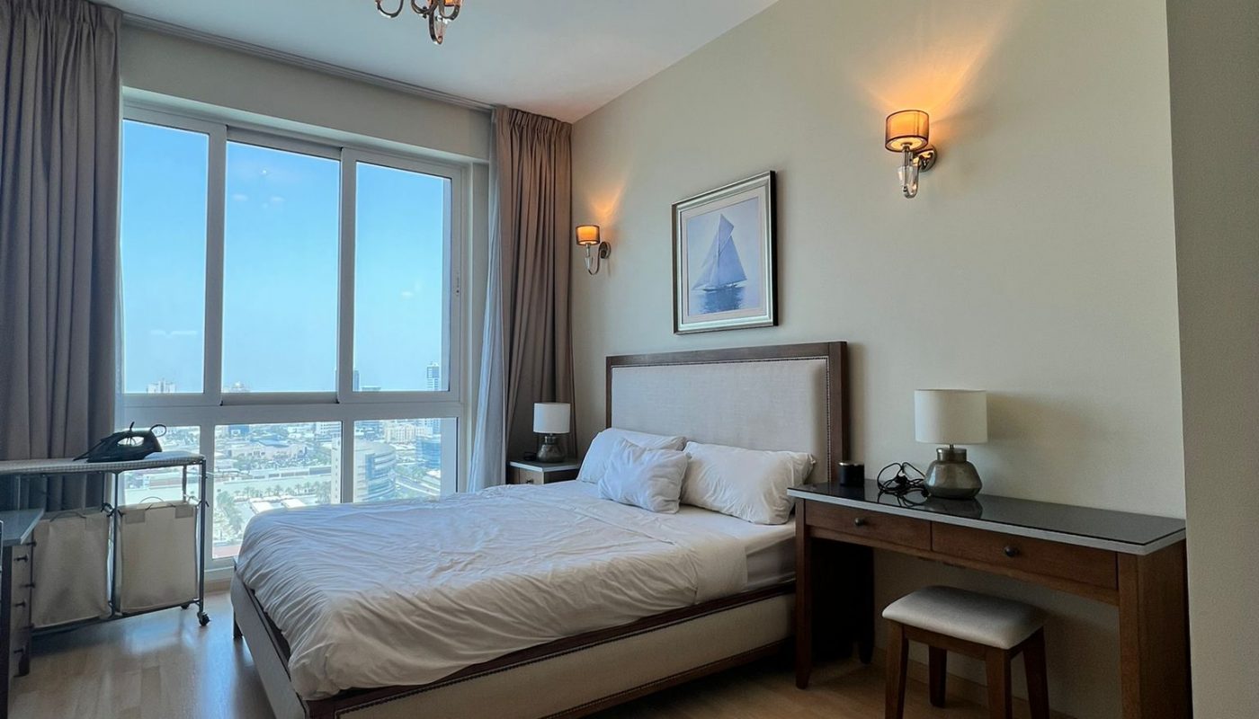A modern bedroom with a large bed, wooden floor, desk, stool, wall art, and large windows with city view, lit by ceiling and wall lamps.