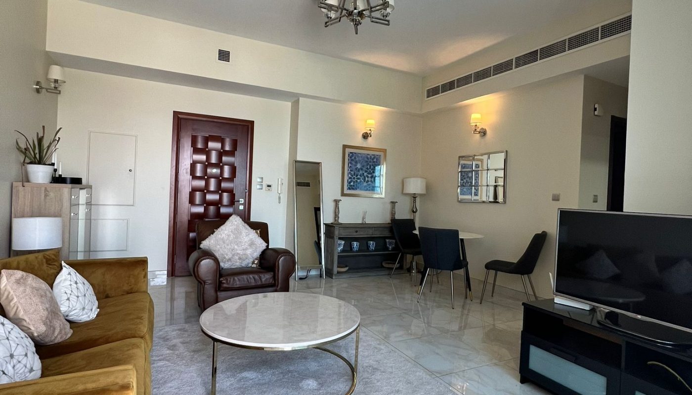 Modern living room with a gold sofa, round coffee table, TV, dining area with two chairs, and neutral decor. Marble floors, light walls, and a central chandelier are visible.