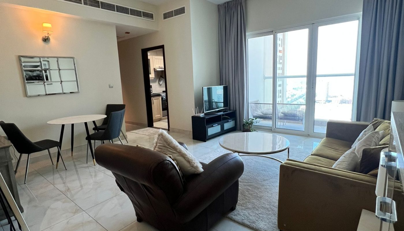 Modern living room with tile flooring, a brown armchair, tan sofa, TV setup, dining area, and large window with balcony and city view.