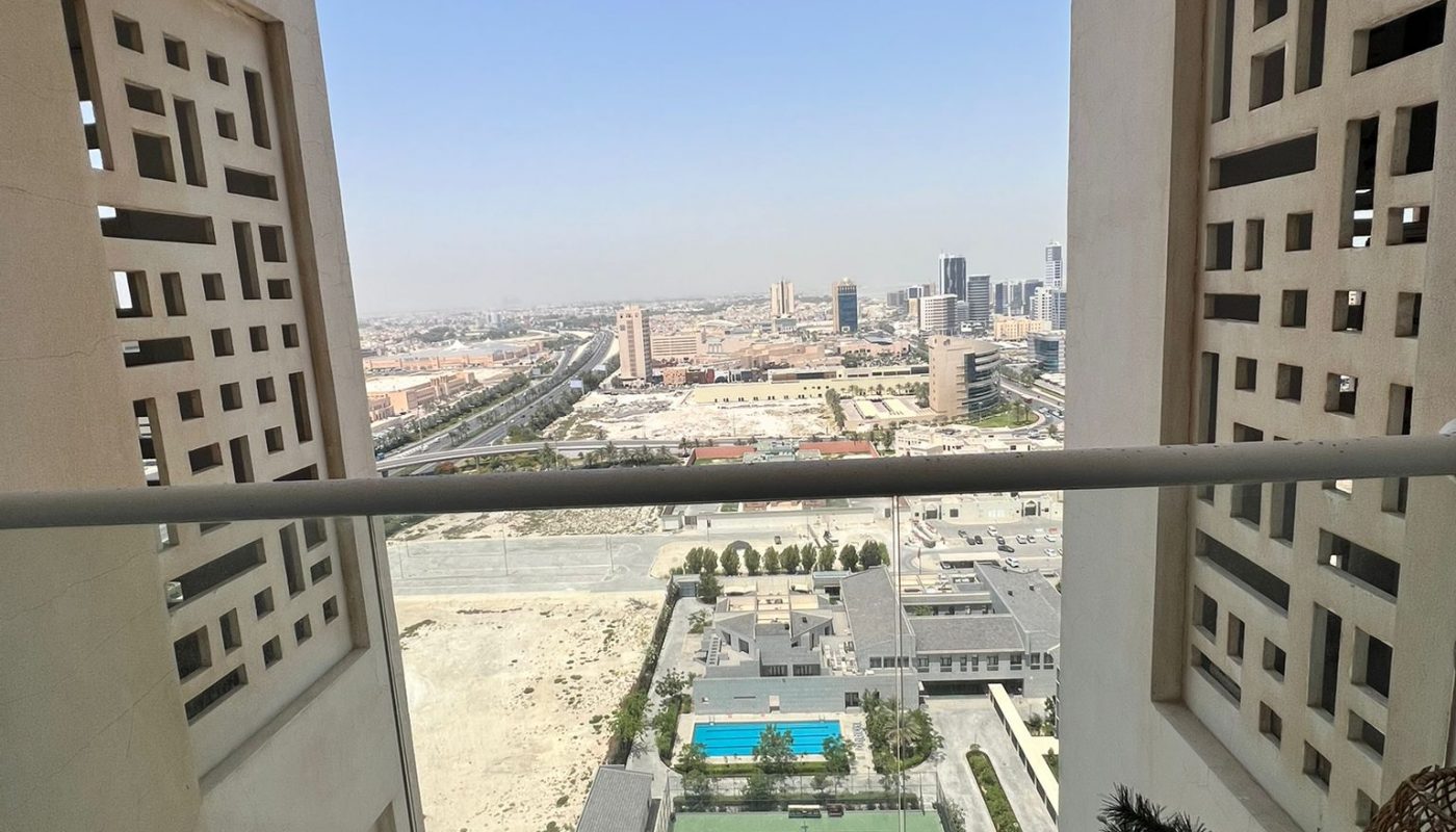 View from a high balcony overlooking a cityscape with buildings, empty lots, a swimming pool, and tennis courts below under a clear sky.