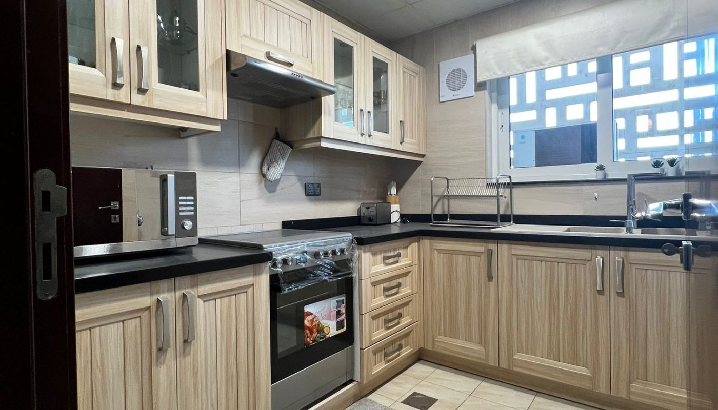 Modern kitchen with light wood cabinets, black countertops, a stove, microwave, toaster, and large window with blinds. A grey rug is on the tiled floor.