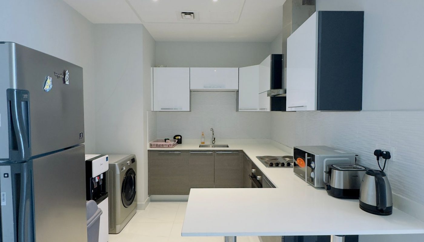 Modern kitchen with white and grey cabinets, stainless steel fridge, washing machine, countertop appliances, and a white breakfast bar.