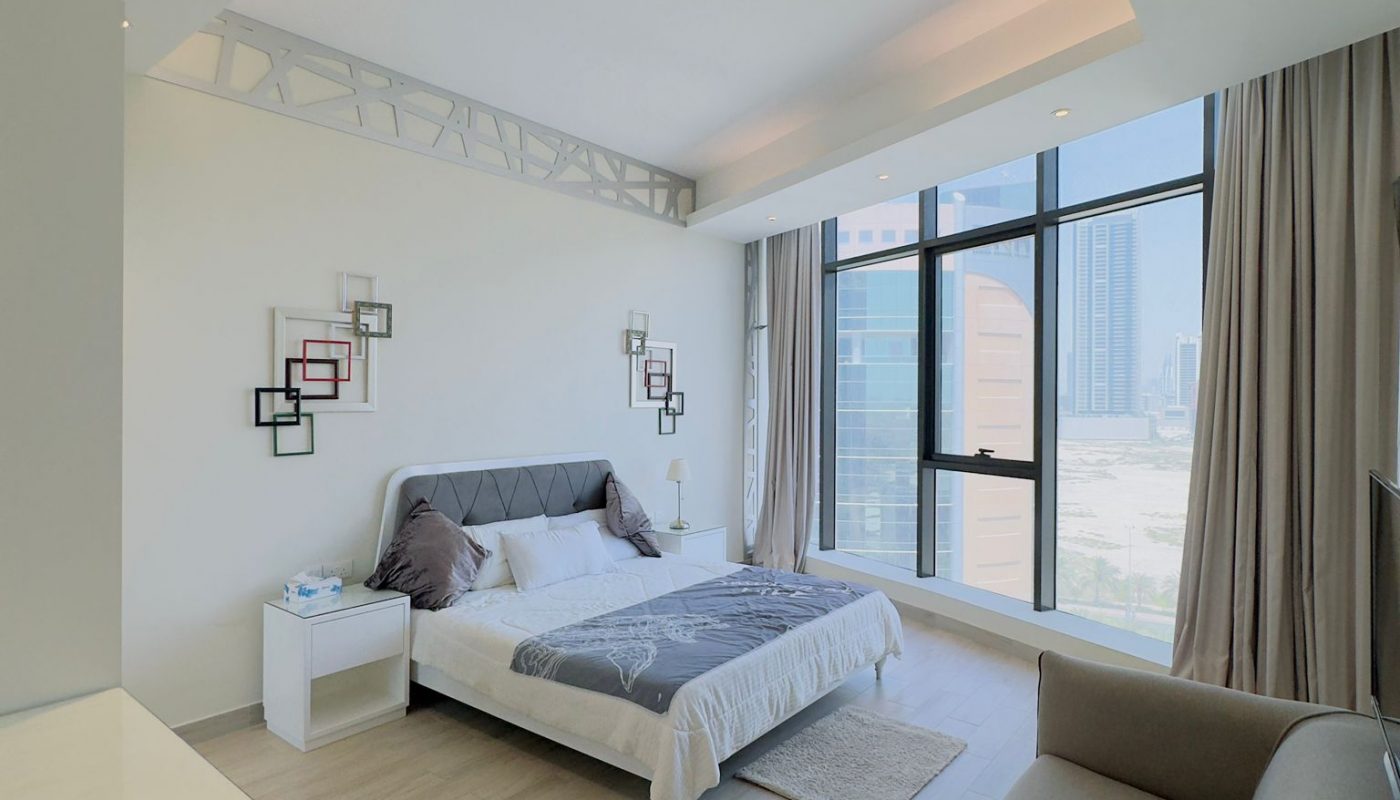 A modern, minimalist bedroom with a large bed, sofa, nightstands, wall art, and floor-to-ceiling windows overlooking city buildings.