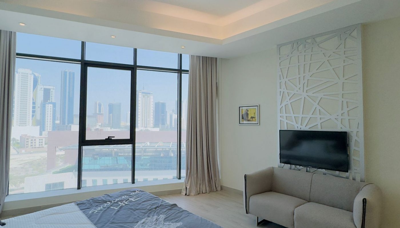 Modern bedroom with large windows overlooking a cityscape, featuring a bed, small sofa, wall-mounted TV, and neutral decor.