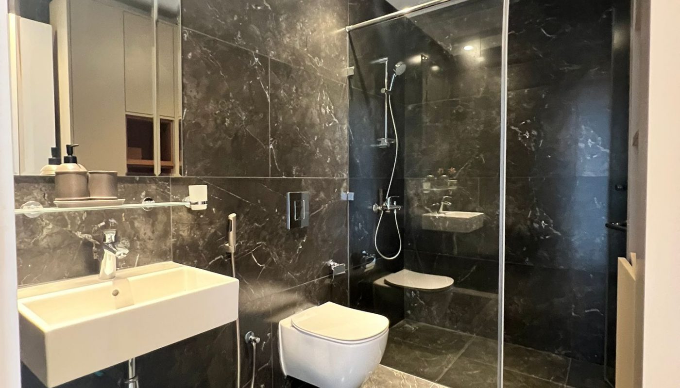 Modern bathroom with dark marble walls and floor, a wall-mounted sink, toilet, handheld bidet, and glass-enclosed shower area. Ceiling has recessed lighting and ventilation.