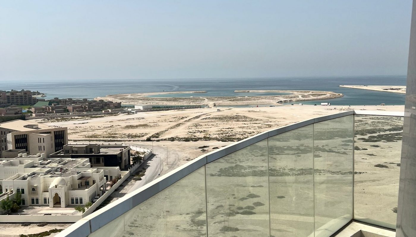 View from a balcony with glass railing overlooking sandy land, buildings, and the sea under a clear blue sky.