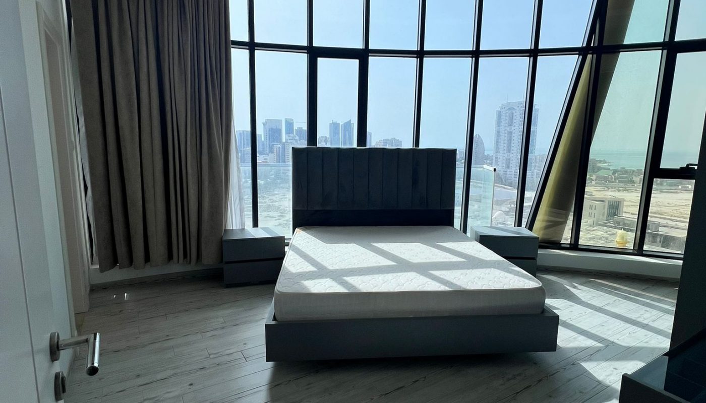 Modern bedroom with a large bed, floor-to-ceiling windows, city skyline view, and light wood flooring.