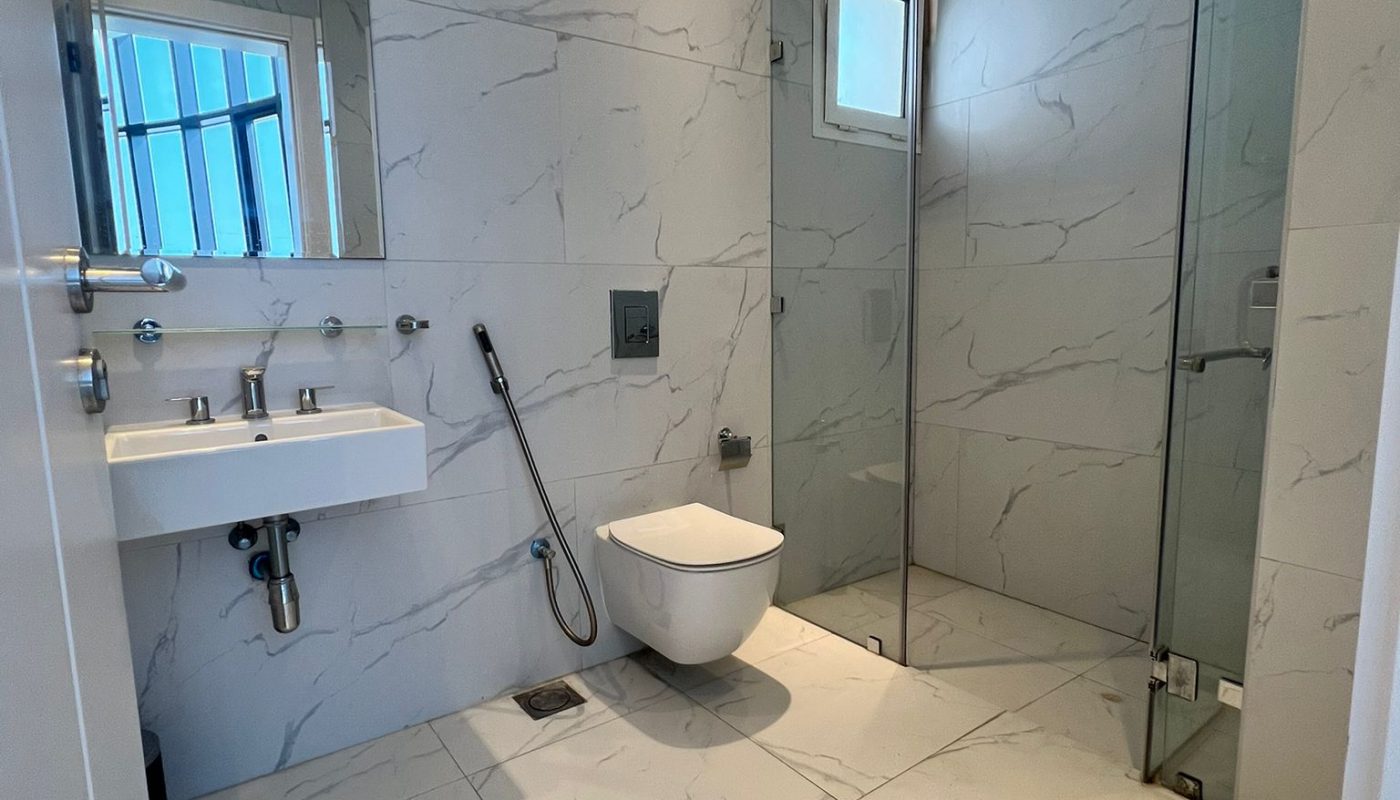 Modern bathroom with marble-patterned tiles, wall-mounted sink and toilet, glass shower enclosure, and a grab bar next to the toilet.