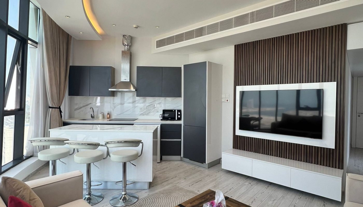 Modern open-plan living room and kitchen with white and gray décor, bar stools, a wooden coffee table, wall-mounted TV, large windows, and neutral furnishings with red accent pillows.