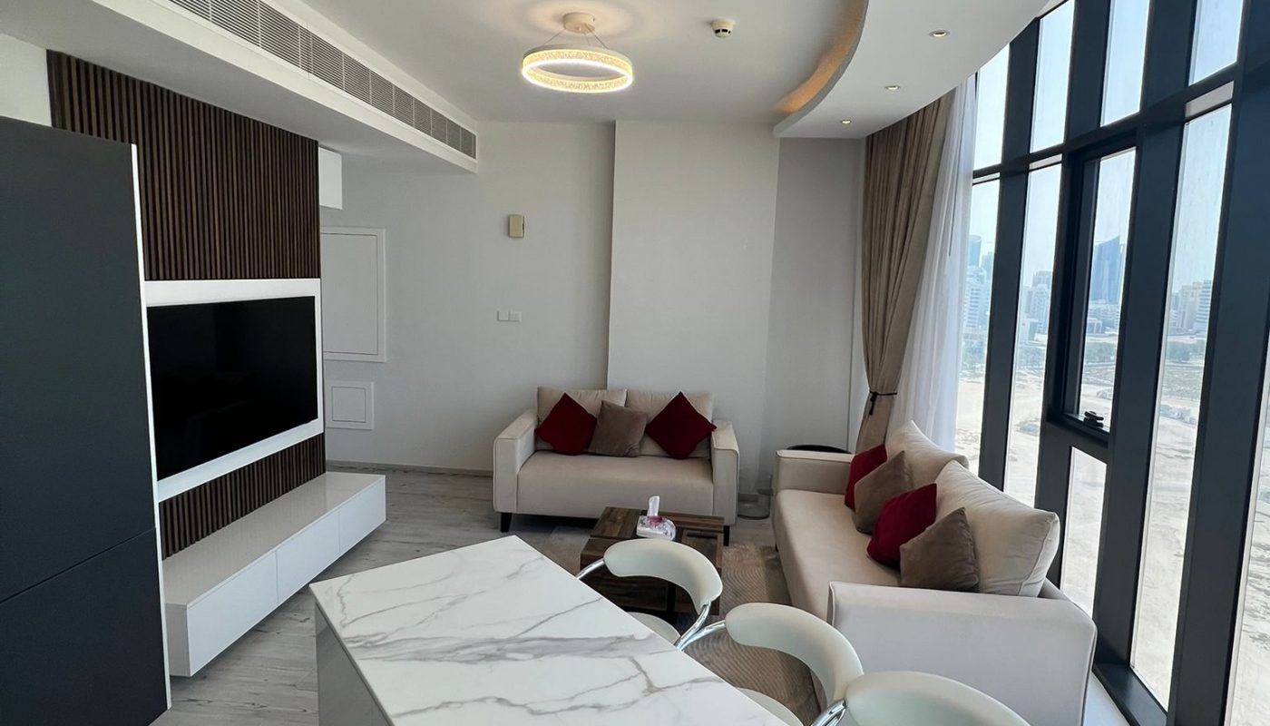 Modern living room with marble kitchen island, two sofas with red pillows, large windows, light wood flooring, and a mounted TV.
