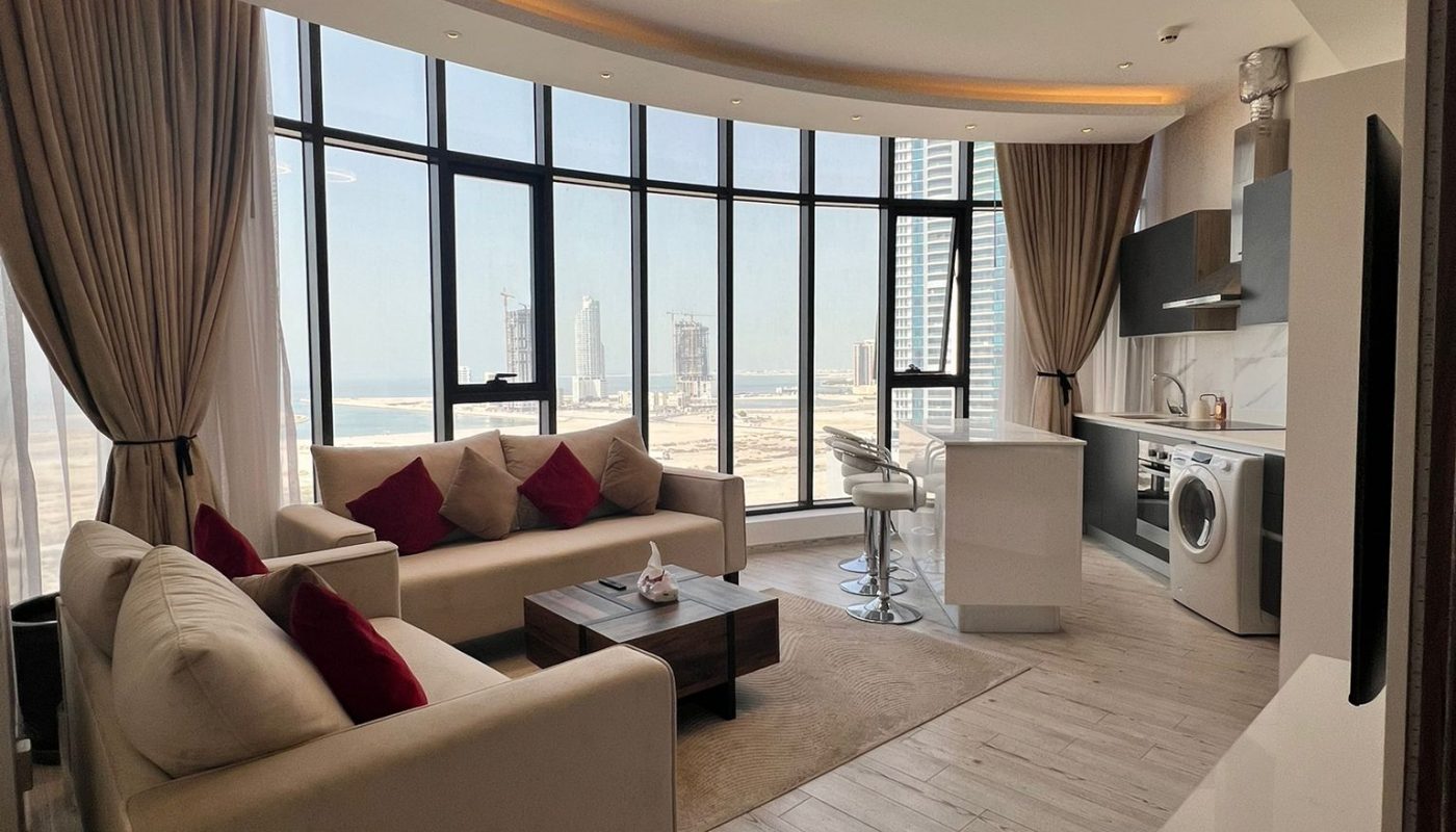 Modern open-plan apartment with large curved windows, beige sofas with red cushions, a kitchen area, and city views in the background.