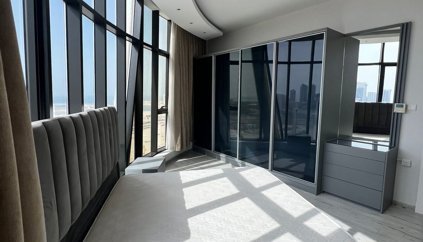 Modern bedroom with large windows, a grey upholstered bedframe, mattress, mirrored wardrobes, and cityscape view; sunlight streams in, casting shadows on white wood flooring.