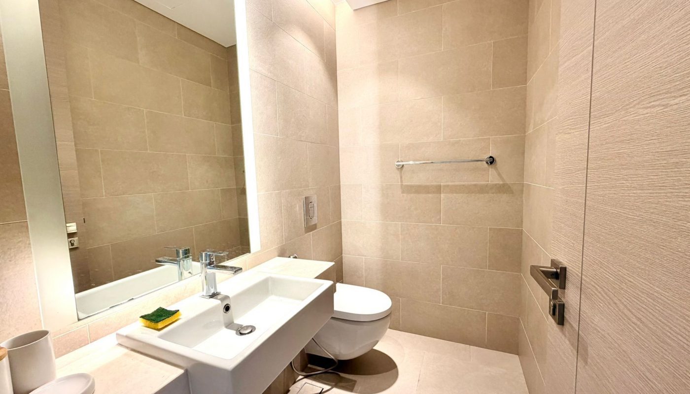 Modern bathroom with beige tiled walls, a wall-mounted sink, a floating toilet, a large mirror, and a towel bar. The room is brightly lit with overhead lighting.