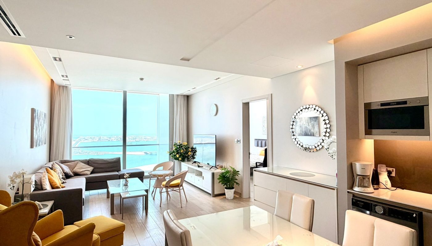 Modern living room and dining area with large window, sea view, yellow armchair, gray sofa, white furniture, and kitchen appliances visible.